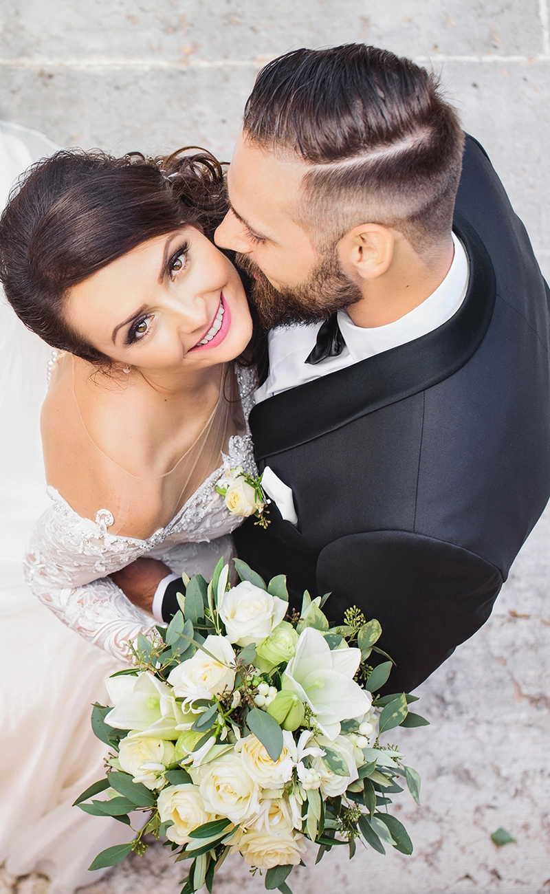Bride with rose bouquet and groom kissing brides cheek while the bride is looking up to the camera over top of them.