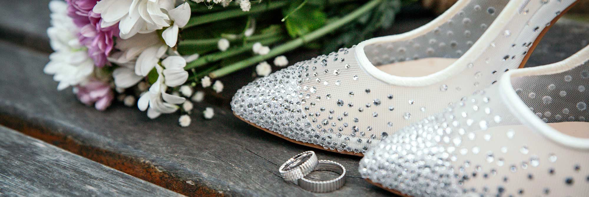 White shoes with rhinestones, with white gold wedding rings and white and pink flowers with babies breath sitting on a grey wood table.<br />
